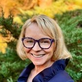 A woman in glasses wearing a blue dress standing in front of a bush.